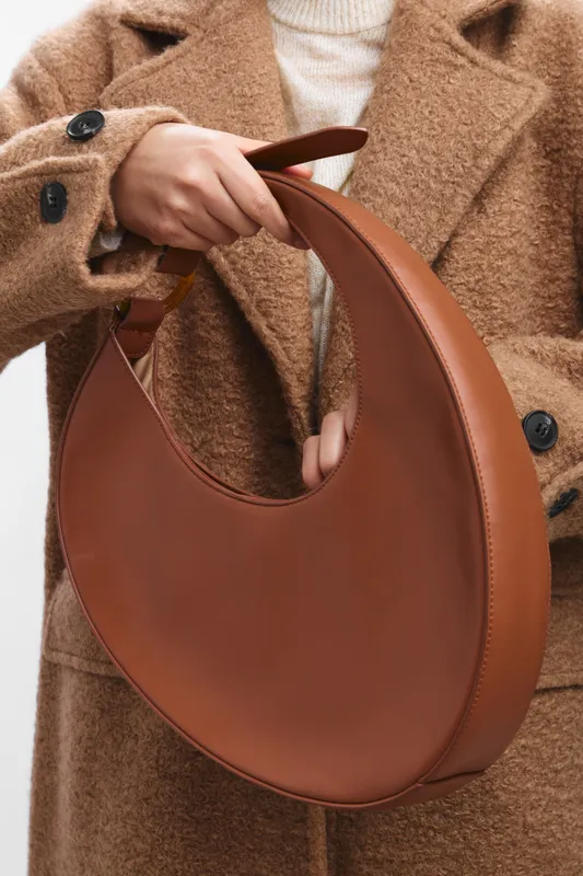 asian woman in a chic beige coat holds a brown circular handbag, showcasing contemporary street fashion with a calm, confident presence and elegant style.