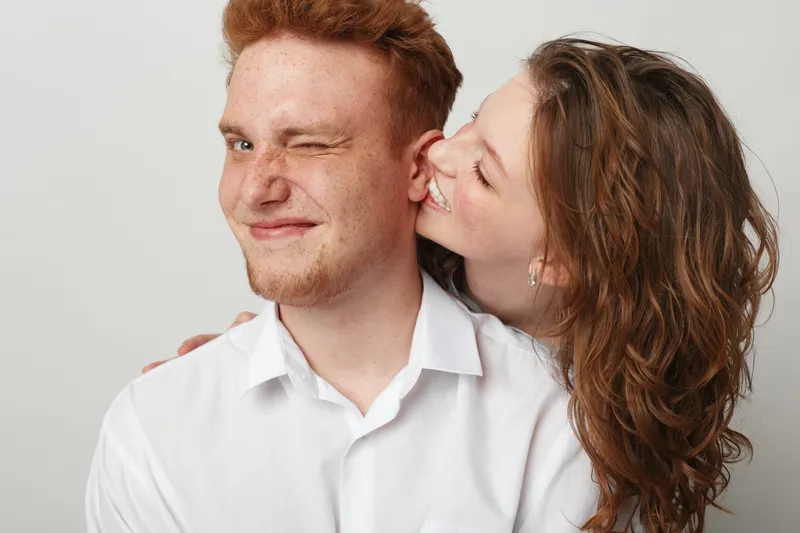 Happy young woman with curly hair kisses smiling man with red hair and beard on cheek. Couple shows affection and joy in casual white shirts on light background. Concept of love and friendship.