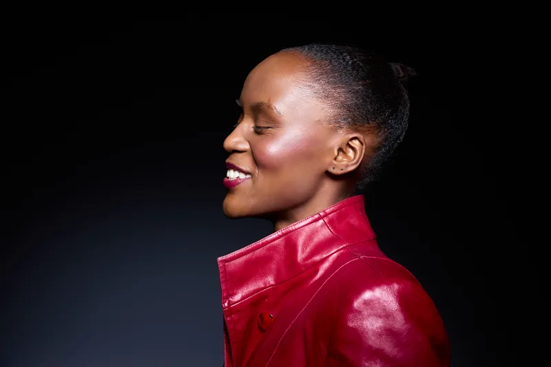 Profile of a smiling African woman with dark skin wearing a red leather jacket against a black background. Concept of confidence, fashion, and natural beauty in studio lighting.