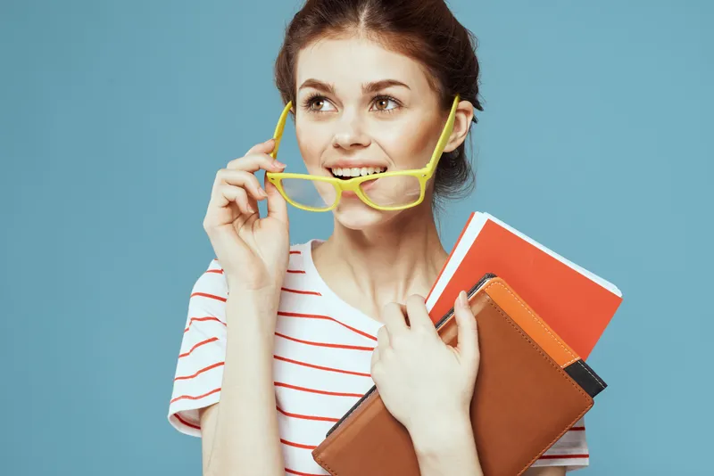 Студентка с книгами в руках на синем фоне, в жёлтых очках и с модельной причёской, кадрированный вид