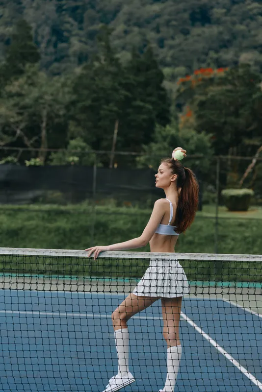 Stylish female tennis player in athletic wear stands confidently on a court, showcasing fitness and determination in a lush green backdrop