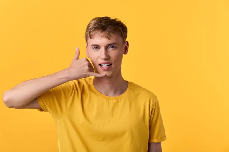 Smiling male in a yellow shirt makes a phone sign gesture against a bright yellow backdrop, conveying casual communication and friendly vibe for lifestyle branding
