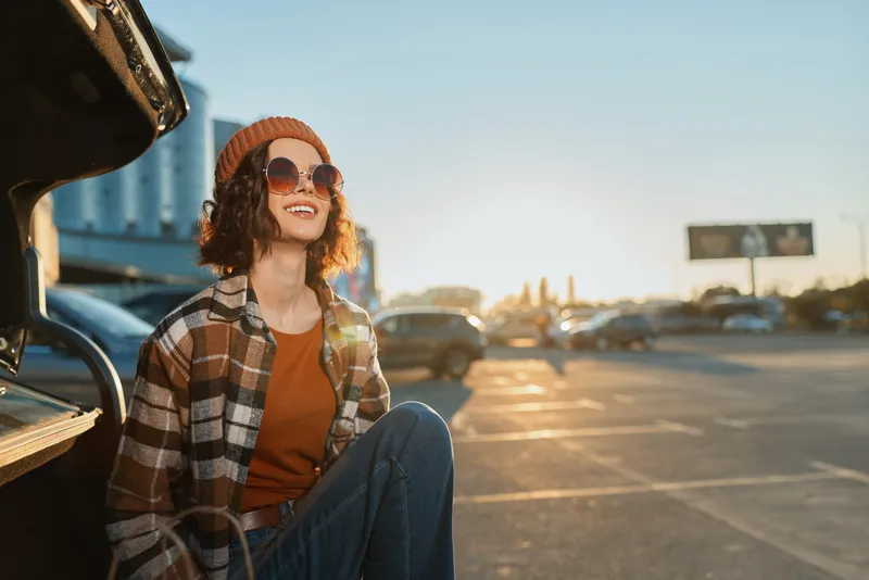 Young woman in beanie and sunglasses leans on a car trunk in a parking lot at sunset, smiling candid, Urban golden hour glow, authenticity, mindful living, emotional storytelling.