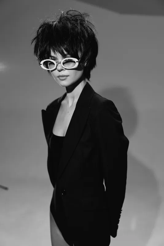 Stylish woman wearing white sunglasses and black blazer posing confidently in black and white.