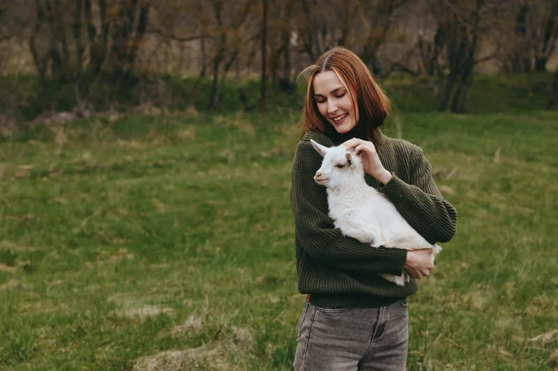 A woman holds a white goat kid in a grassy field, smiling in a cozy knit sweater, capturing a serene outdoor lifestyle moment in a countryside setting, calm and affectionate