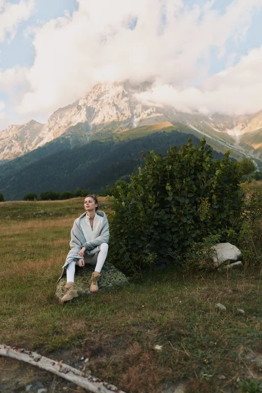 Woman lifestyle portrait sits on grass beside a shrub with distant mountains, calm reflective mood, outdoor scene, natural light, tranquil moment
