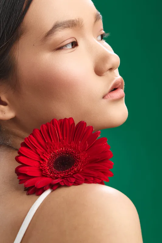 asian woman is captured in a close portrait with a red flower resting on her shoulder, soft lighting highlights delicate features, serene expression, and a graceful, artistic mood.