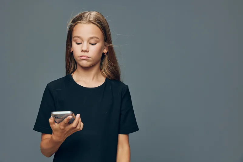 Girl with long hair in black shirt looking at phone with serious expression on gray background for technology, social media, youth and communication concept.
