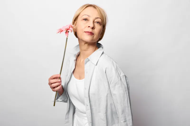 Mature woman holding a pink flower in studio portrait, wearing white blouse and light jacket, calm thoughtful expression, soft minimal grey background, neutral copy space.