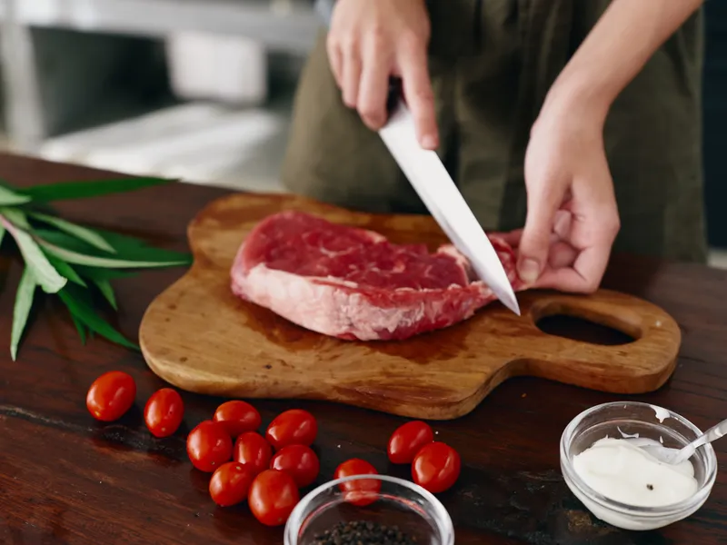 Женщина с ножом в руке нарезает свежее стейковое мясо для жарки на кухне