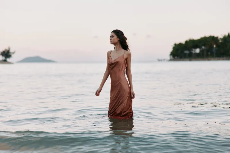A woman stands waist-deep in calm sea water, wearing a rust-colored slip dress, gazing toward the horizon as soft evening light fades on the shore.
