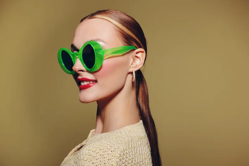 Stylish woman with long hair wearing large green sunglasses and red lipstick poses against beige background. Concept of fashion, beauty, and modern accessories.