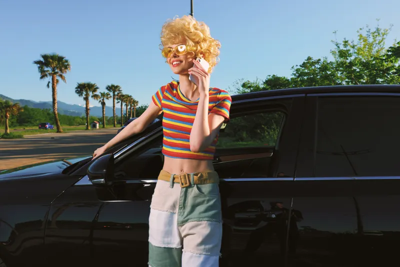 Young woman with curly blonde hair leans on a sleek black car in a sunny desert setting wearing a striped crop top and patchwork skirt radiating retro confidence.