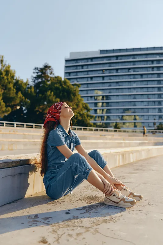 lifestyle woman in modern boho-western denim street style wearing red bandana relaxing outdoors in film color with urban background and casual sneakers
