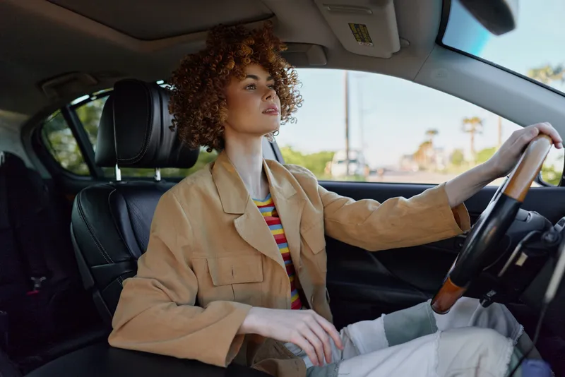 Young woman with curly hair drives a stylish car, wearing a tan jacket and striped shirt, radiating focus and calm as she navigates the road.