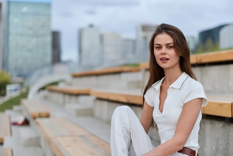 Young woman with thoughtful expression in casual white outfit sitting on urban outdoor benches with cityscape background, natural light, fashion lifestyle portrait