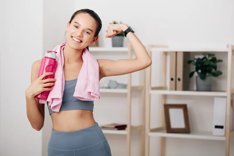 Woman in athletic wear flexing her bicep with a pink towel and water bottle in a bright home gym, showcasing fitness, health and workout motivation.