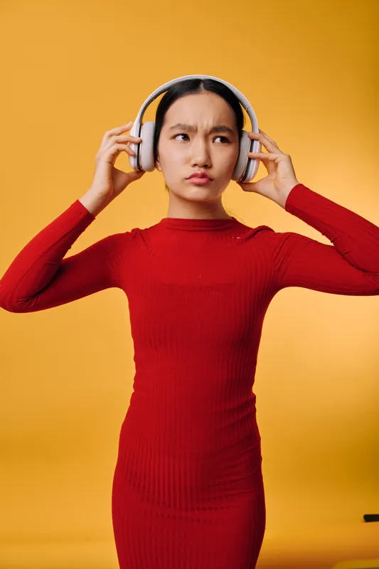 Young woman in red dress wearing white headphones with curious expression listening to music on colorful yellow background studio. Female person standing isolated with modern device for audio