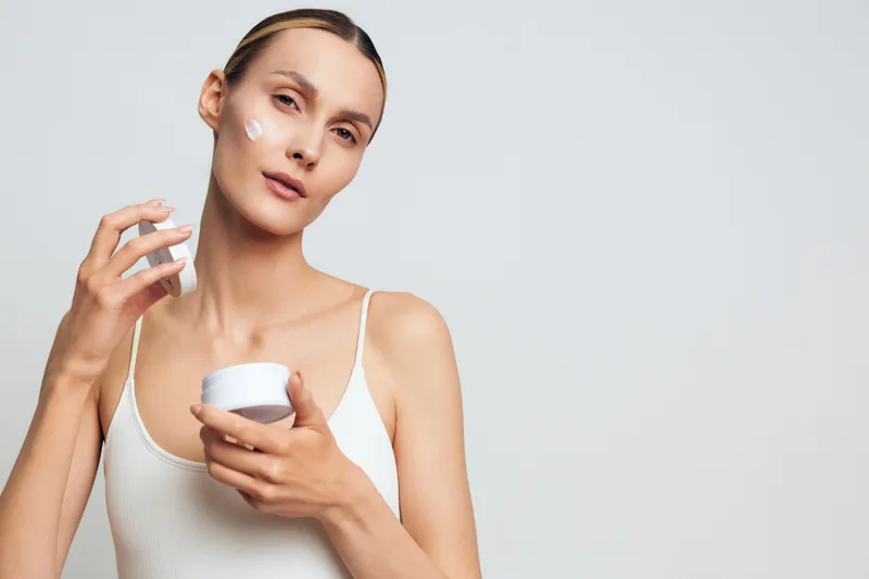 Young woman applying white face cream on her cheek with a calm expression on a light background. Concept of skincare, beauty routine, self-care, and healthy skin.