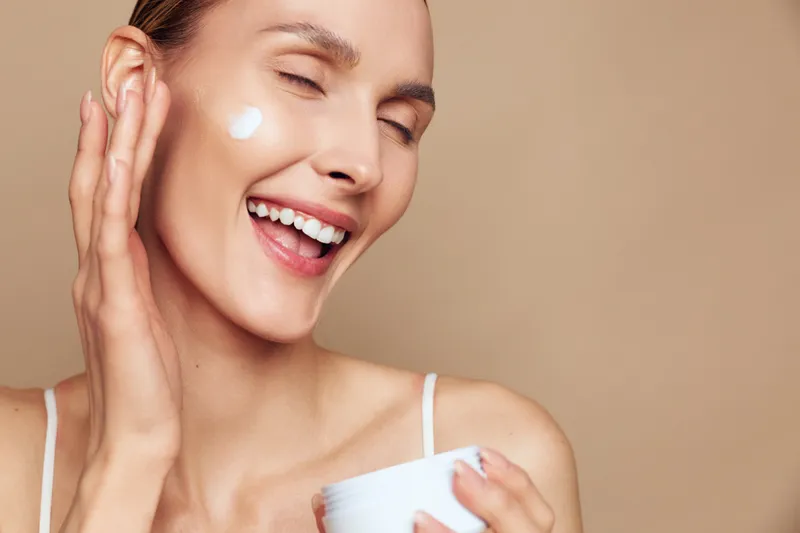 Happy young woman with clean skin applies face cream on cheek smiling with closed eyes isolated on beige background enjoying skincare routine and natural beauty care.