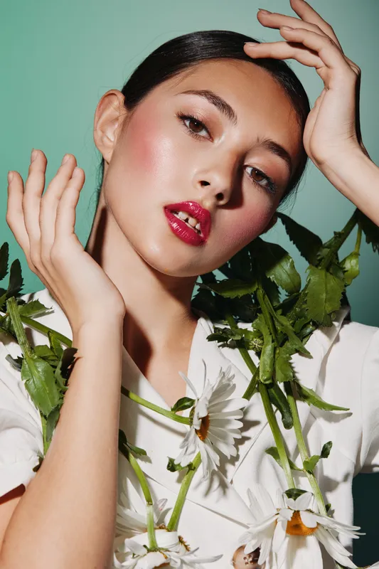 Close-up of a young woman with glossy red lipstick wearing a white floral blouse, fresh makeup highlights, and natural lighting against a mint green backdrop for fashion and beauty concept.
