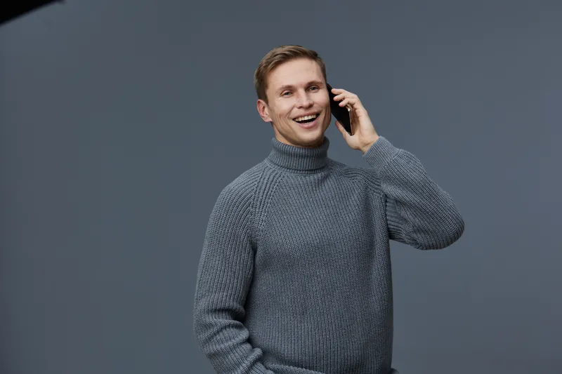 Smiling man talking on mobile phone, casual grey turtleneck sweater, cheerful expression, indoor studio shot, modern lifestyle, bright mood, friendly atmosphere, confident young adult male, isolated b