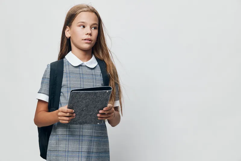 Schoolgirl with backpack holding tablet looking sideways in studio with copy space for education technology digital learning and student concept on white background.