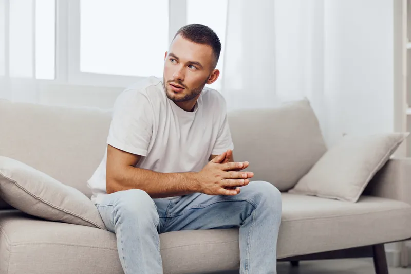 Thoughtful man sitting on a cozy couch, wearing casual attire, reflecting on life and future. Bright, airy atmosphere emphasizes a sense of introspection and calmness.