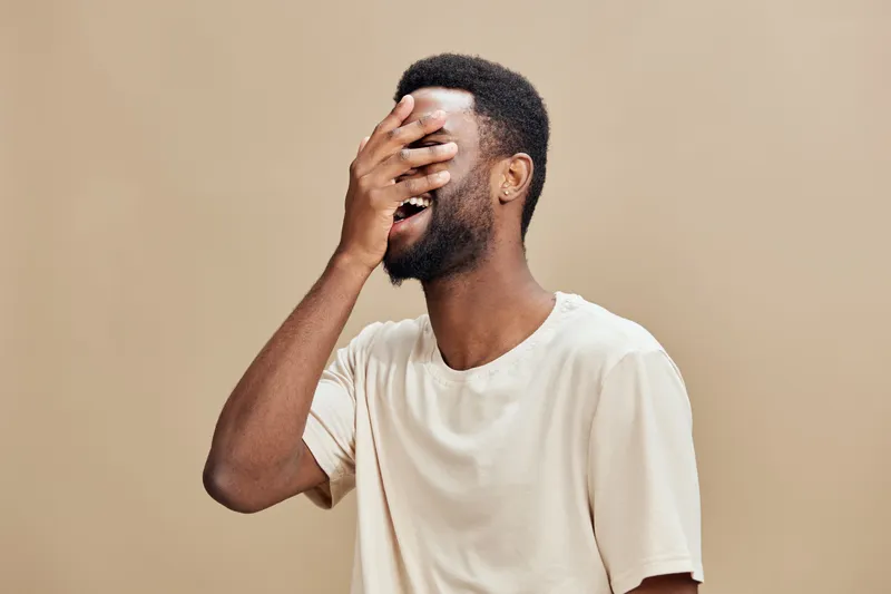 Happy young African man laughing while covering his face with hand, isolated on beige background, showcasing joy and emotion in a modern style