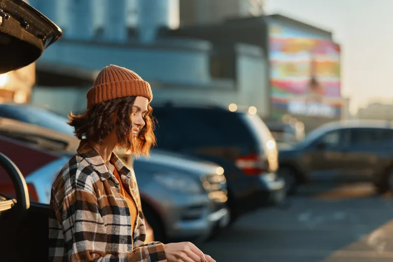 woman smile car parking urban sunlight beanie candid lifestyle happiness portrait outdoors golden hour glow. Young woman in plaid coat by car trunk at sunset showing authenticity, mindful living and
