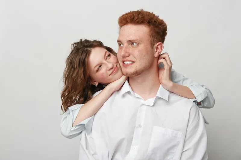 Young couple in white shirts embraces and smiles against a plain background. The woman hugs the man from behind, showing affection and happiness in a casual moment.