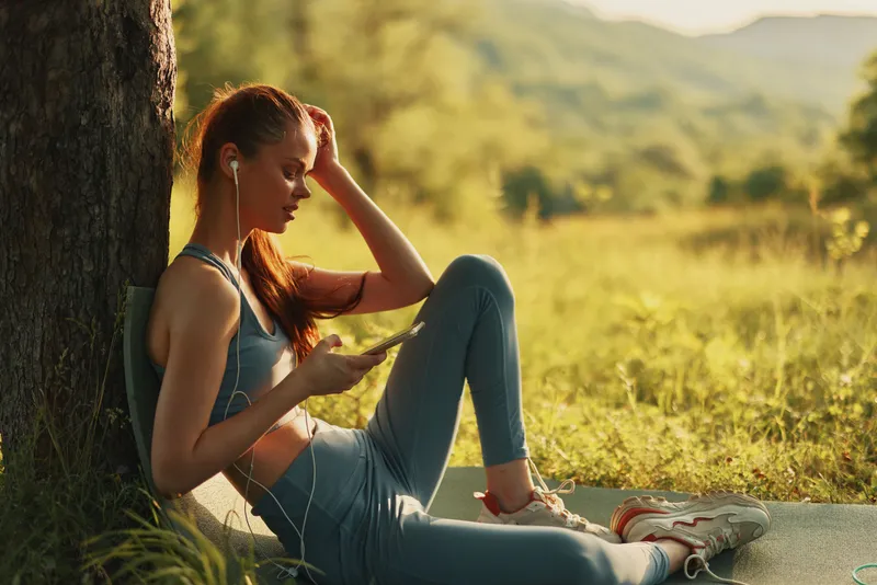 Young woman in sportswear relaxing outdoors, listening to music, enjoying nature in a peaceful summer setting, representing wellness and leisure