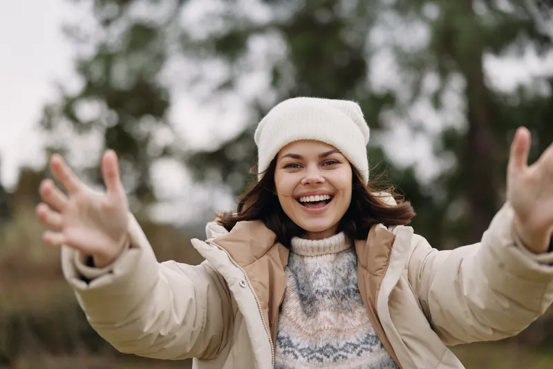 Woman outdoors in winter attire with a warm jacket and beanie, smiling with arms open, expressing joy and freedom in a chilly landscape