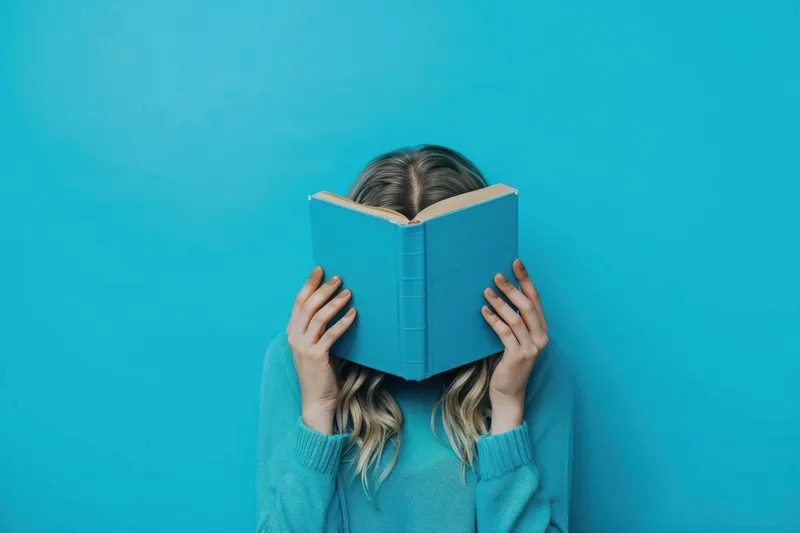 Женщина читает книгу и закрывает ею лицо на синем фоне в спокойной обстановке