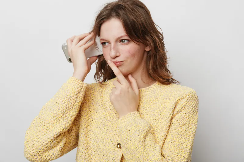 Young woman in yellow sweater holds smartphone to her ear and looks thoughtful, finger on lips. Concept for communication, decision making, and contemplation in casual setting.