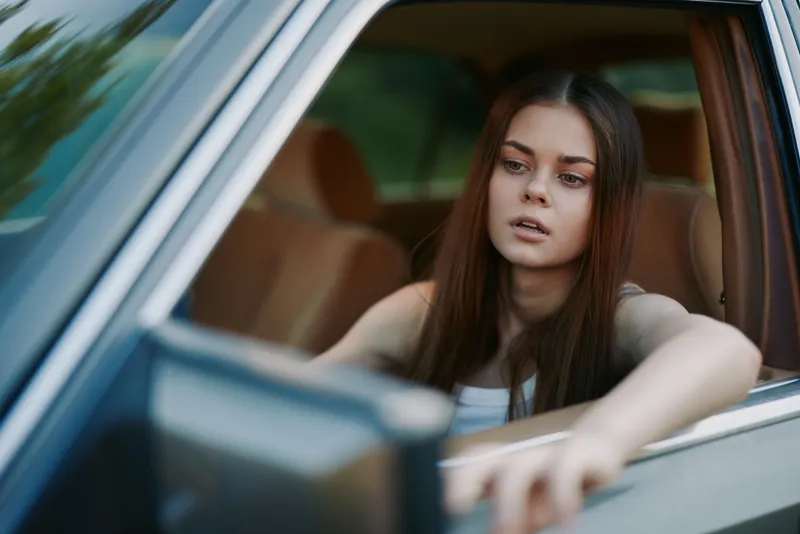 Thoughtful young woman inside a car, with long hair and casual attire, expressing contemplation against a blurred green background.
