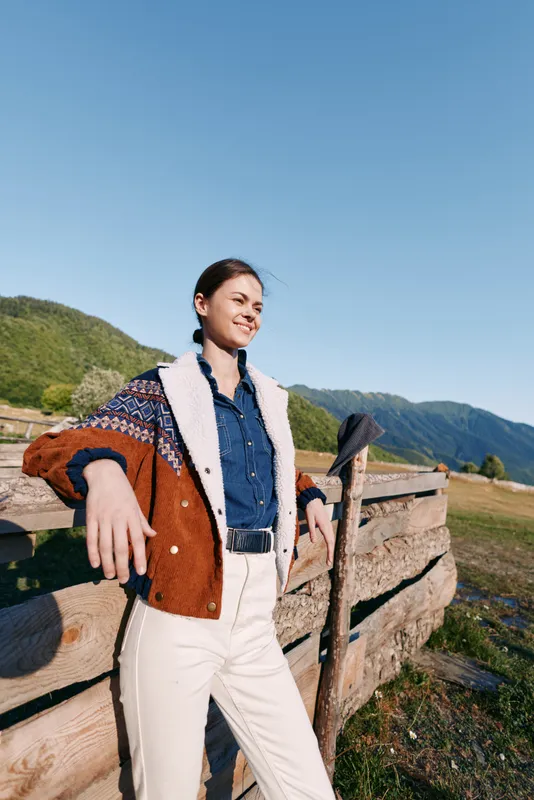Woman in countryside leaning on wooden fence with mountains behind, outdoors portrait showing smile and relaxed casual cardigan style against blue sky and green meadow.