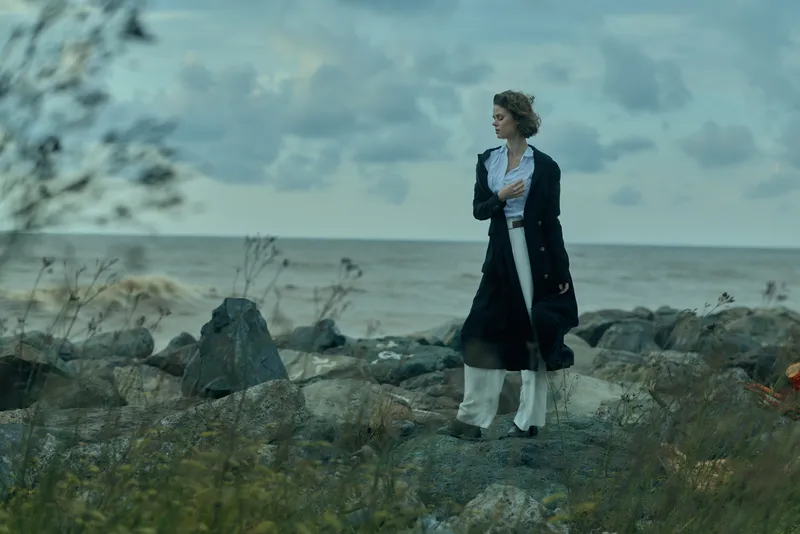 Woman in a dark overcoat and white dress stands on a rocky shoreline by the sea, a windswept contemplative moment with moody clouds and rugged coast