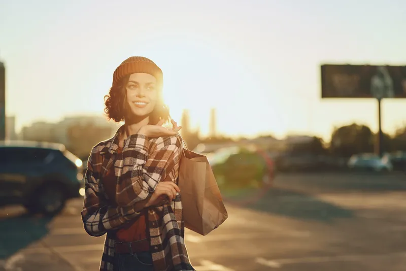woman smile shopping golden hour urban candid lifestyle authenticity: Young woman carries a paper shopping bag across a city street at sunset, joyful expression, warm golden hour glow, mindful living