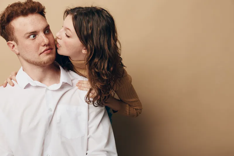 Young woman kisses surprised man on cheek while hugging him from behind. Casual couple in warm beige studio background. Concept of affection, love, and playful relationship moment.
