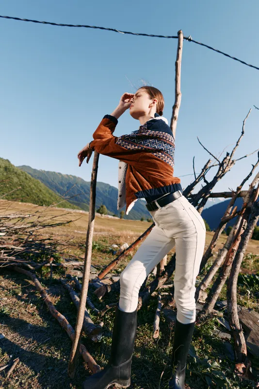 Woman model in countryside wearing a cozy sweater and boots, striking a stylish pose by a wooden fence with mountains and blue sky in background, outdoor fashion portrait.