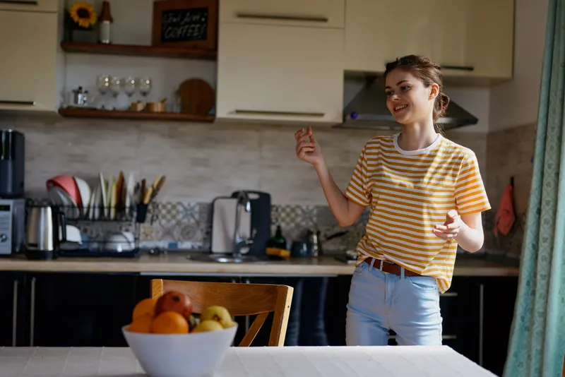 A woman in a striped yellow shirt smiles and gestures in a bright modern kitchen, standing beside a fruit bowl. A cozy home lifestyle scene with everyday positivity and domestic warmth.