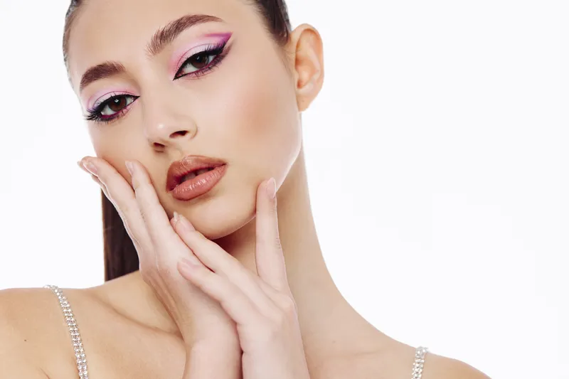 Young woman with dramatic pink eyeliner and glossy lips poses with her hands near her face in a creative beauty fashion studio shot on a white background.