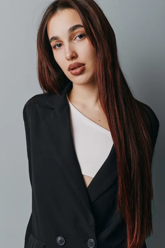 Young woman with long straight hair wearing black blazer and white top posing against gray background. Fashion portrait of confident female model with natural makeup and serious expression.