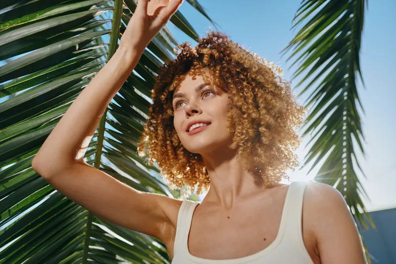Smiling woman with curly hair enjoys sunlight outdoors near tropical palm leaves, wearing white tank top, bright natural light, blue sky background.