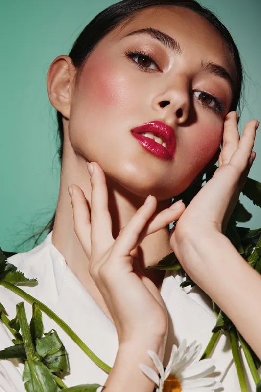 Close-up of a fashionable woman with bold makeup posing elegantly surrounded by green leaves and flowers. Concept of beauty, creative fashion, and natural style.