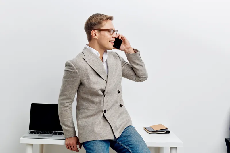 Modern smiling man in smart casual attire talking on mobile phone while leaning on desk with laptop, notebook, and glasses, in bright office environment. People lifestyle concept, professional work, c
