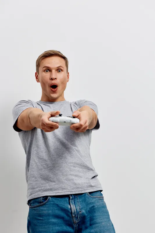 Smiling man playing video game expressing excitement and surprise with a game controller, casual grey t-shirt and jeans, standing against plain white background. Lifestyle entertainment concept for ga