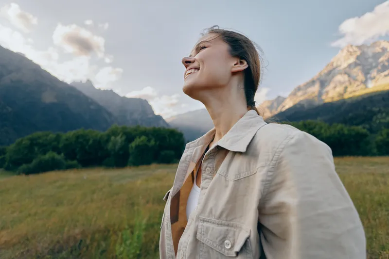 A young woman in a beige jacket stands in a sunlit mountain meadow, tilting her head back with a bright smile, enjoying the fresh air and wide open sky.