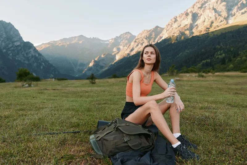 A young woman sits on a grassy field in a mountainous landscape, wearing an orange top and shorts. She gazes into the distance, resting near a backpack and a water bottle.